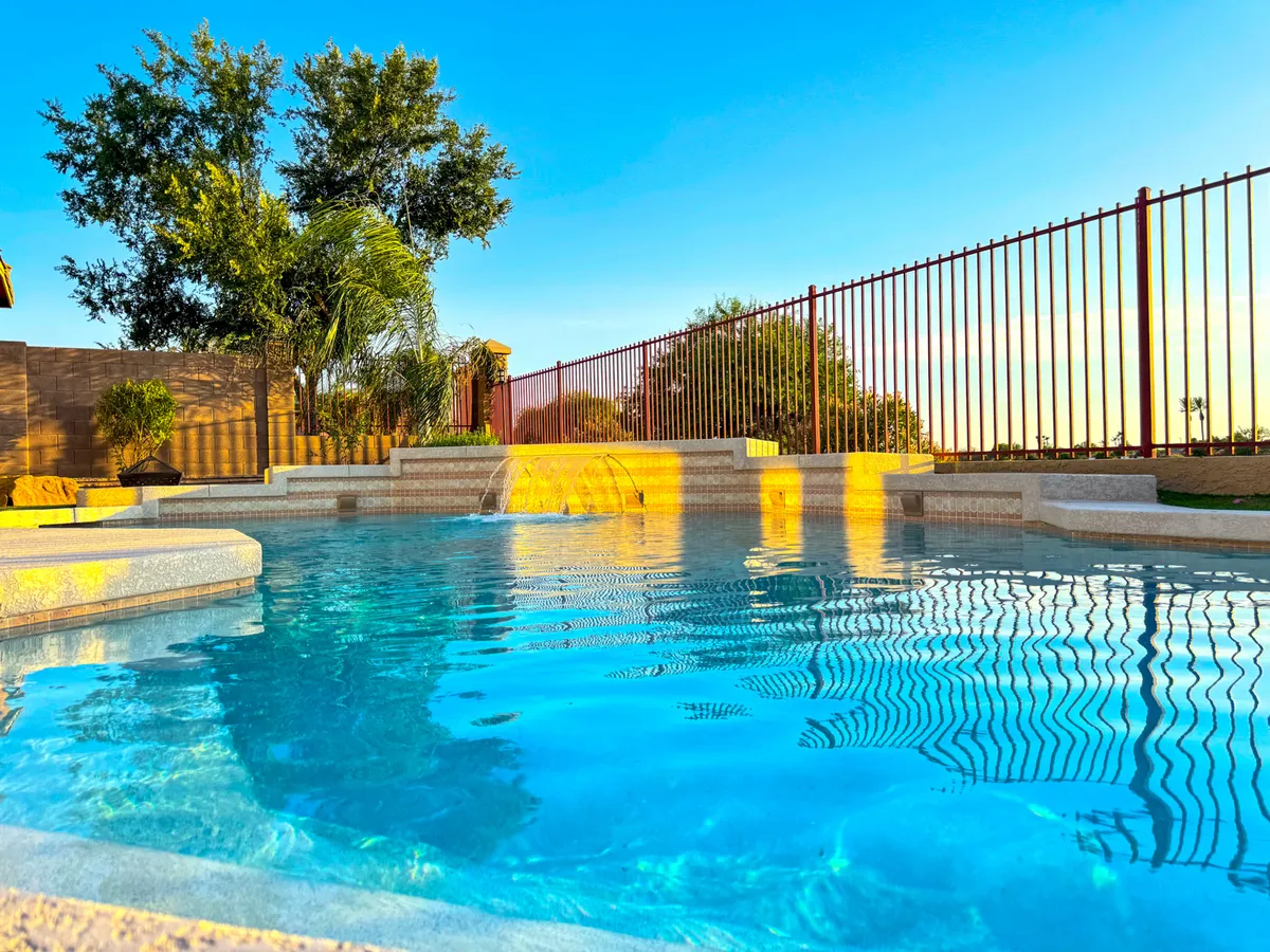 A serene backyard oasis featuring a swimming pool with a waterfall feature, surrounded by lush greenery and a privacy fence. The setting feels refreshing and inviting, suitable for morning swims, family gatherings, or quiet relaxation by the water.