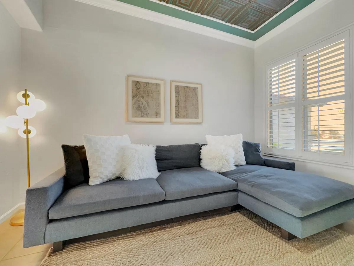 A cozy, modern office or flex space with a stylish sofa, decorative pillows, artwork, and a lamp beside a window. The space feels adaptable—suitable for work, study, or an extra sleeping area.