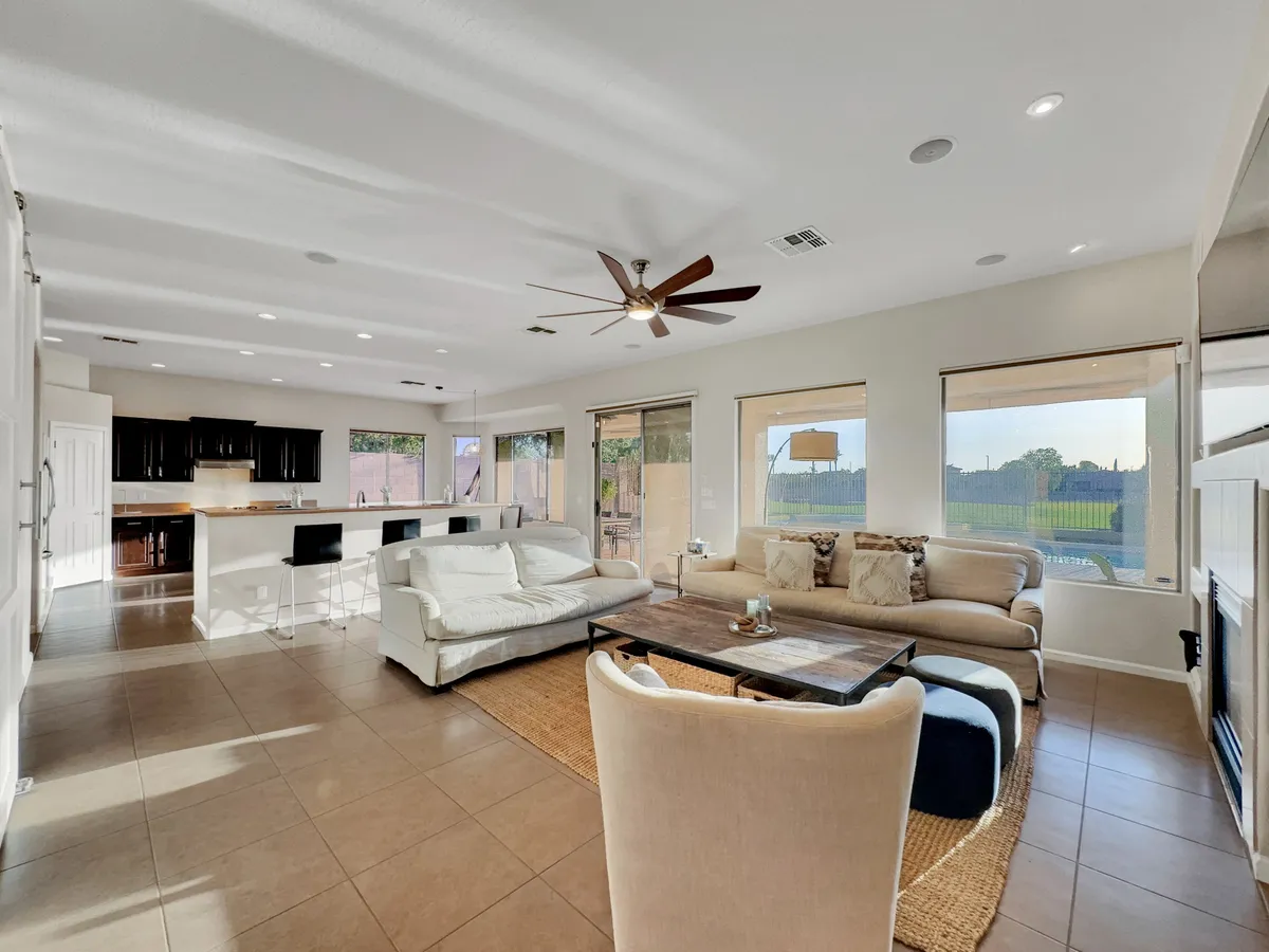 This image features a spacious great room that integrates a large sectional sofa and a wooden coffee table with a kitchen island and bar stools in the background. Ample windows allow natural light to flood the space, and the layout encourages interaction and easy flow between cooking, dining, and relaxing zones.