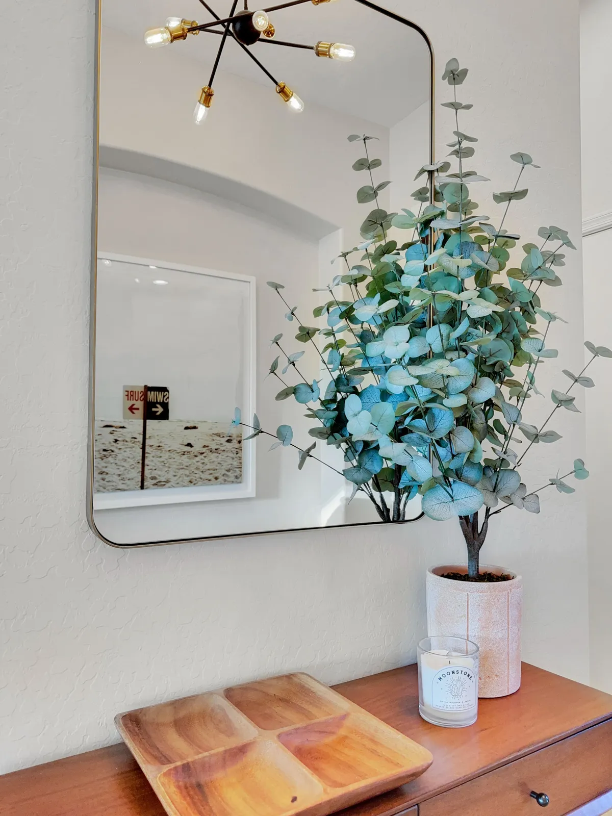 This image features a modern, light-filled entryway with a stylish eucalyptus plant, mirror, and wooden serving tray on a console table. The decor is fresh and inviting, capturing the welcoming spirit as you step into the home.