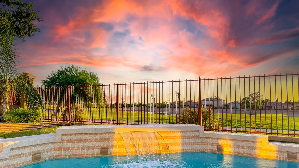 An exterior view of the pool with a stunning water feature, set against a colorful sunset sky. The fenced area and surrounding greenery add to the sense of privacy and relaxation, highlighting outdoor enjoyment during pleasant weather.
