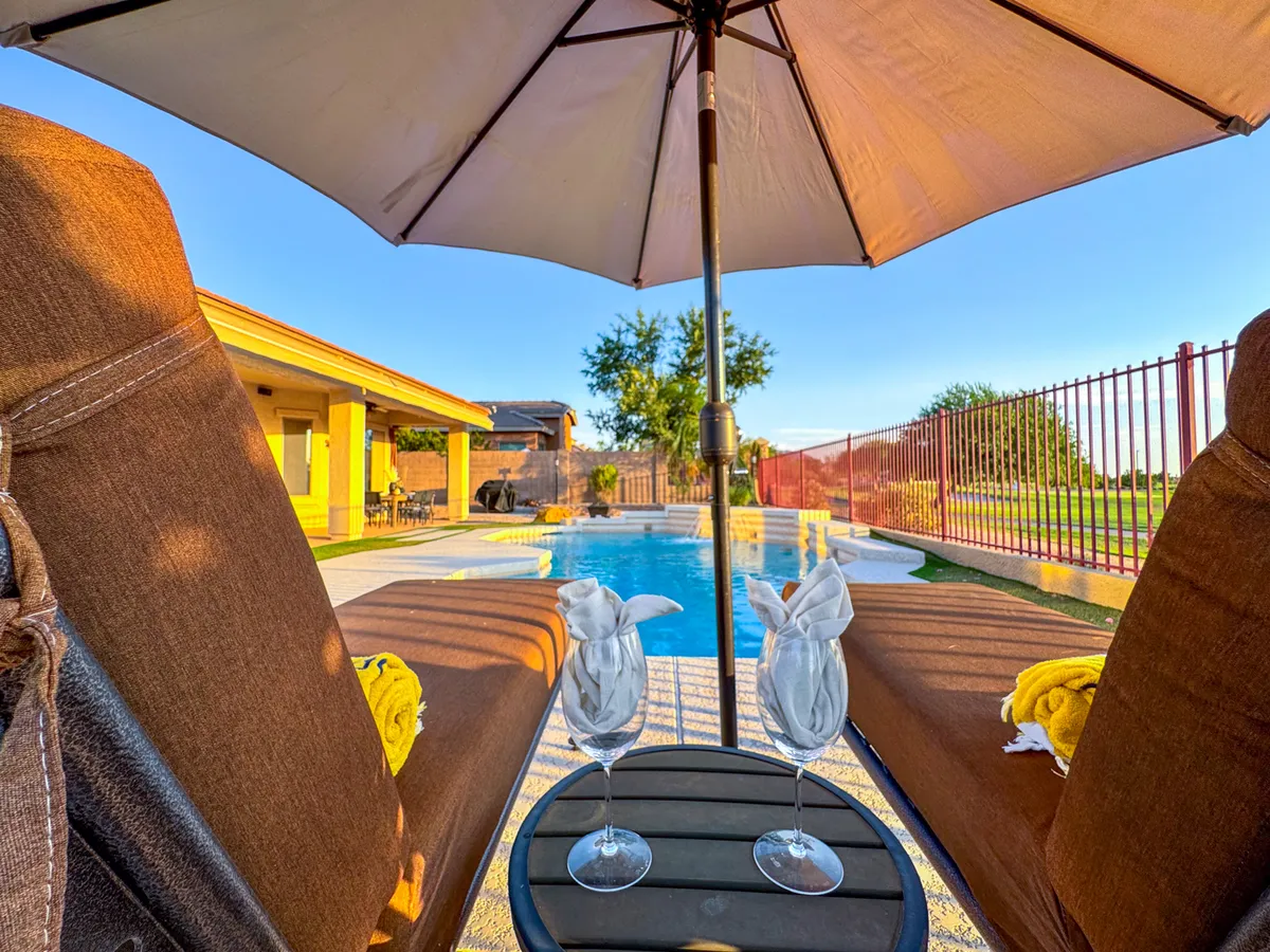 This image showcases a tranquil backyard pool area, complete with inviting lounge chairs, an umbrella for shade, and drinks set out for relaxation. The pool is the focal point, framed by lush greenery and a peaceful atmosphere ideal for enjoying both sunny and cooler days.
