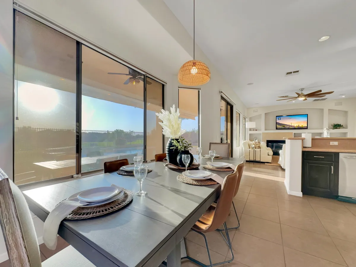 This image captures a stylish dining area with a table set for six, flanked by large windows that let in abundant daylight and provide scenic views. The kitchen area is visible, with a decorative vase with flowers adding a touch of warmth and festivity.