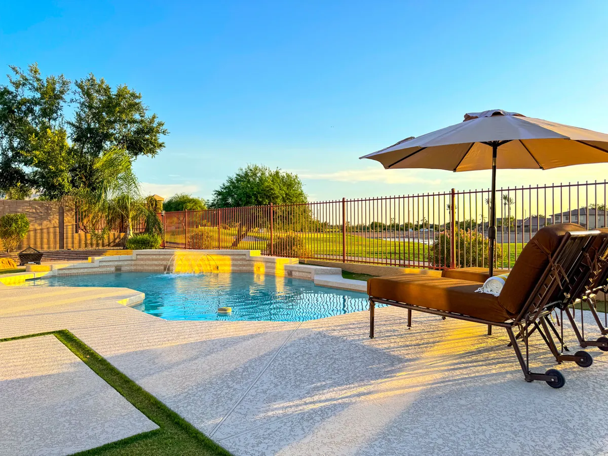 A bright outdoor pool area with lounge chairs, an umbrella for shade, lush landscaping, and a secure fence. The inviting pool is ideal for swimming or sunbathing, while the setup encourages outdoor dining, relaxation, and socializing under the sun or stars.