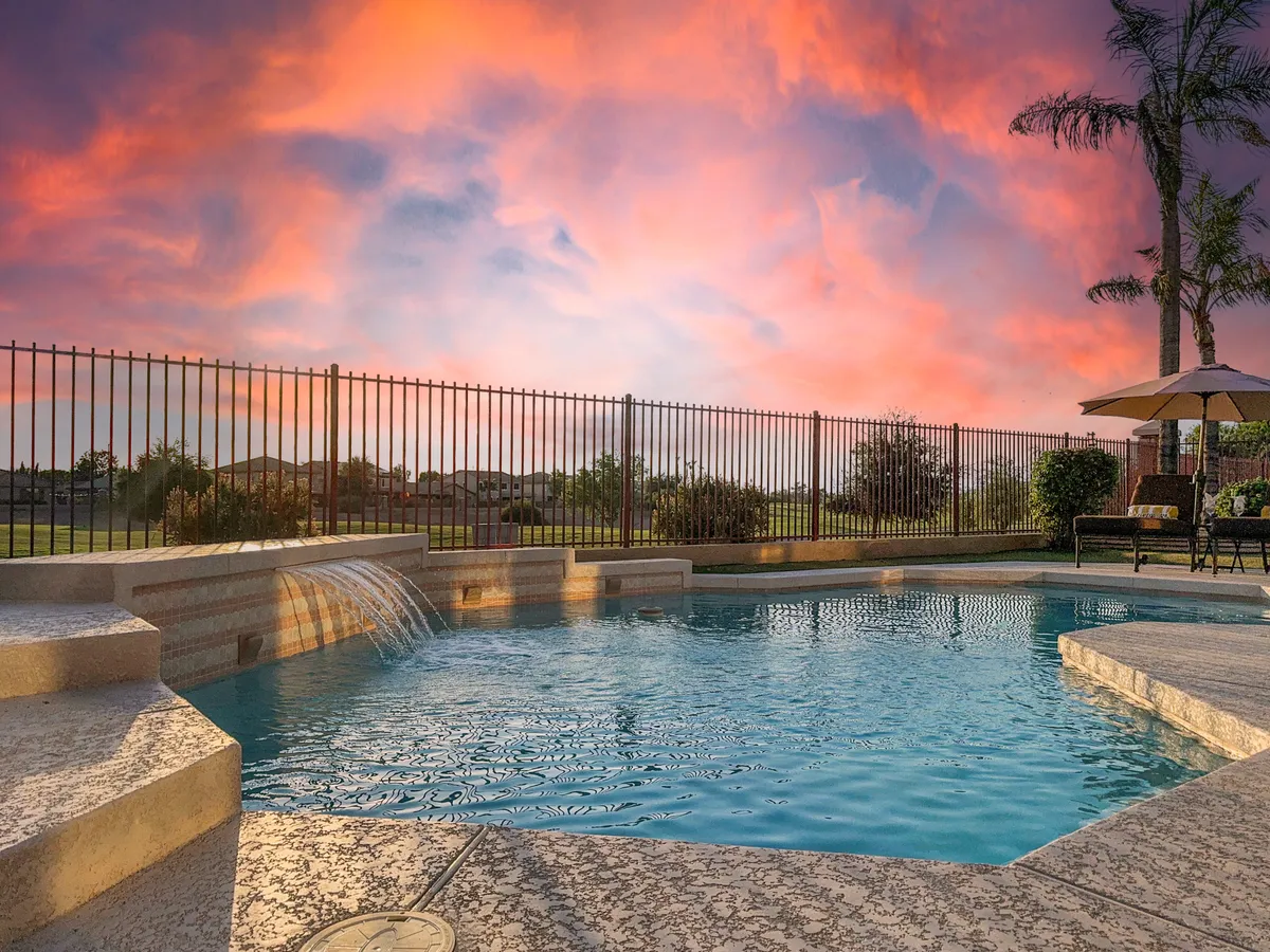 A tranquil backyard scene featuring a sparkling swimming pool with a waterfall feature, surrounded by palm trees and set against a vivid sunset sky. The poolside area offers ample space for gatherings, sunbathing, and relaxation, evoking a resort-like atmosphere.