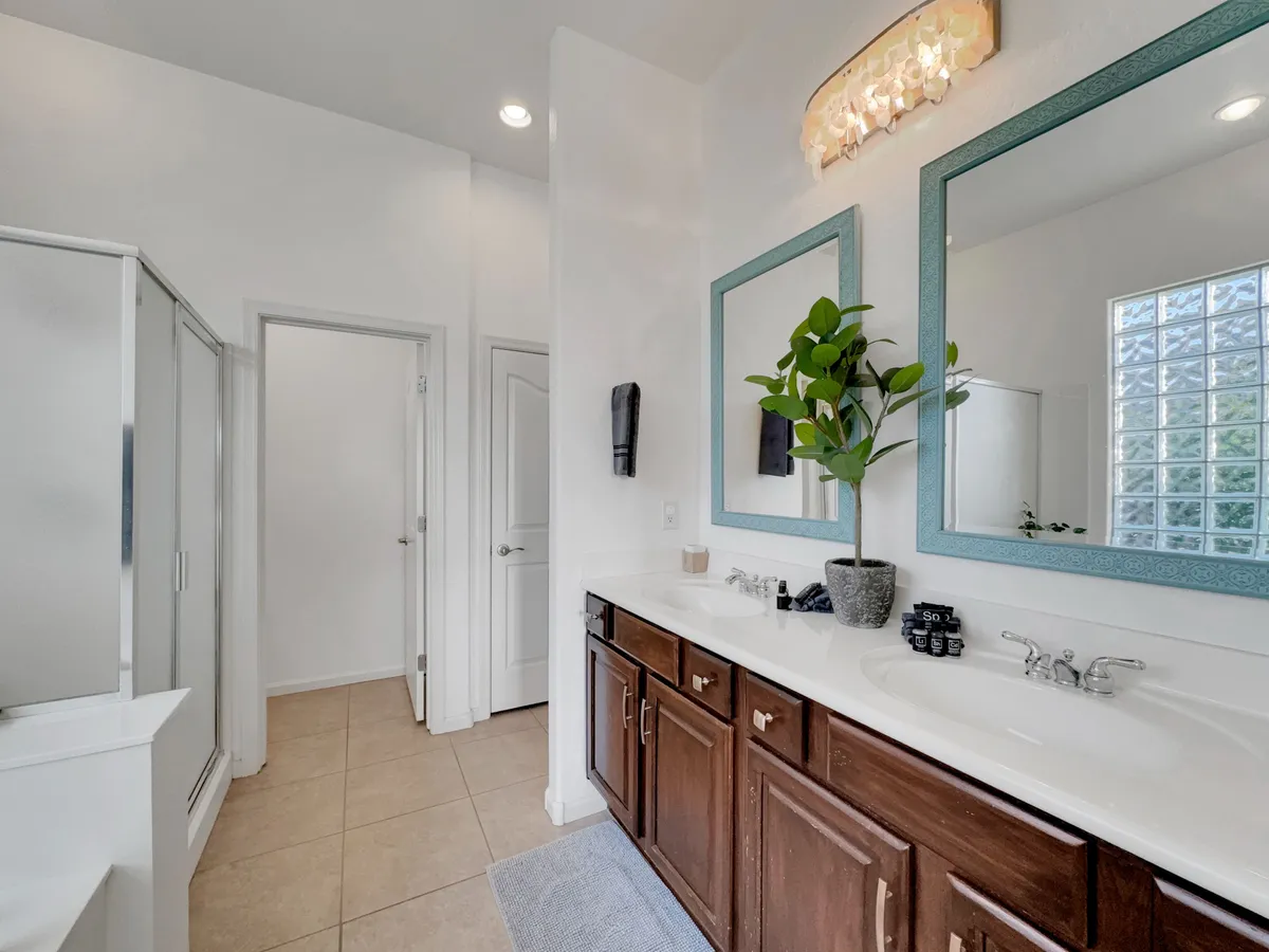 A spa-inspired bathroom with double sinks, mirrors, a glass shower enclosure, and a lush green plant. Natural light pours in from a window, illuminating the calming, spacious space.