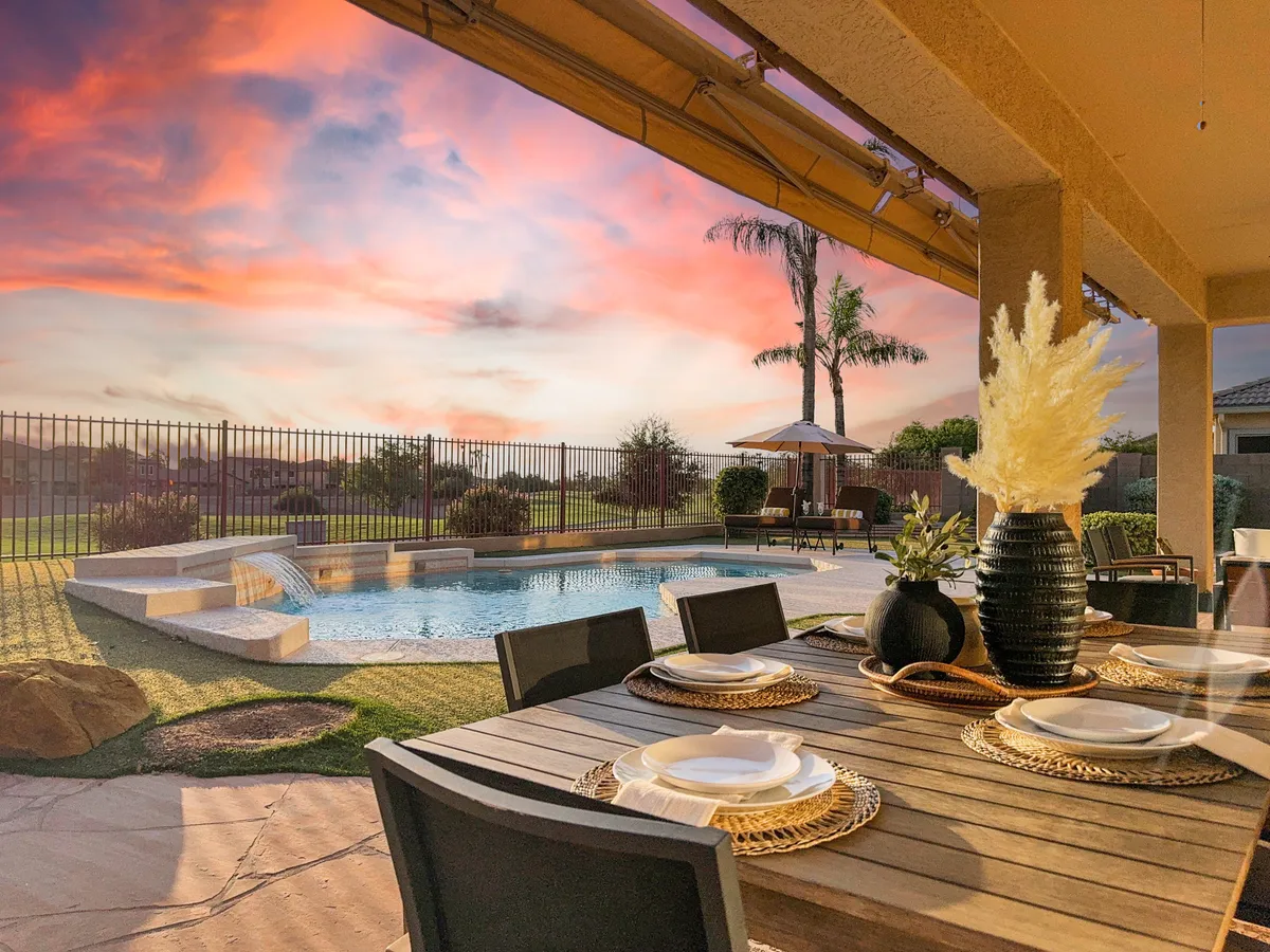 An inviting backyard scene with a set dining table, a glimmering swimming pool, and a vibrant sunset sky. Decorative plants and tableware add elegance, while the pool and seating area exude a relaxed, resort-like atmosphere. The space is clearly designed for both swimming and outdoor dining, with lush landscaping framing the scene.