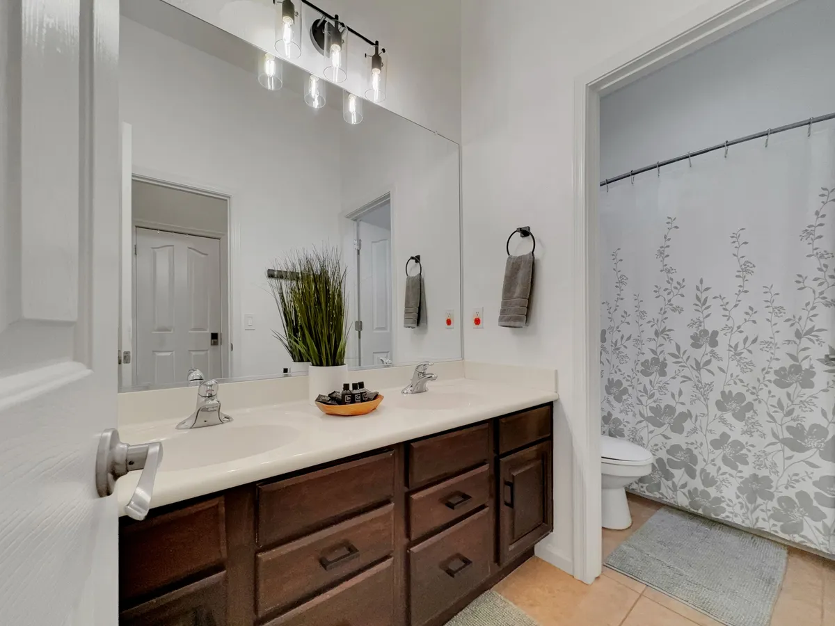 A calm and organized bathroom vanity with double sinks, a large mirror, decorative plant, and towel hooks on the wall. The neutral palette and tidy setup evoke a sense of order and luxury.
