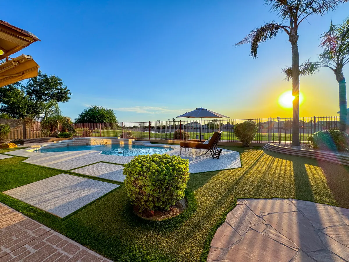 A tranquil scene of a swimming pool surrounded by palm trees, a lounge chair, and a meticulously landscaped garden. The sunset casts a soft glow over the water, inviting relaxation and sunbathing. The setting highlights both the leisure and the natural beauty of the backyard environment.