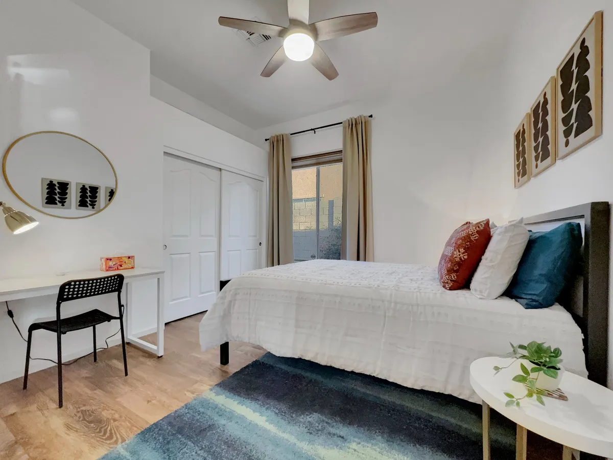 A serene bedroom with a queen-sized bed dressed in soft decorative pillows, a desk with a mirror for work or study, and curtains that let in natural light. The space feels calm and adaptable for both rest and productivity.