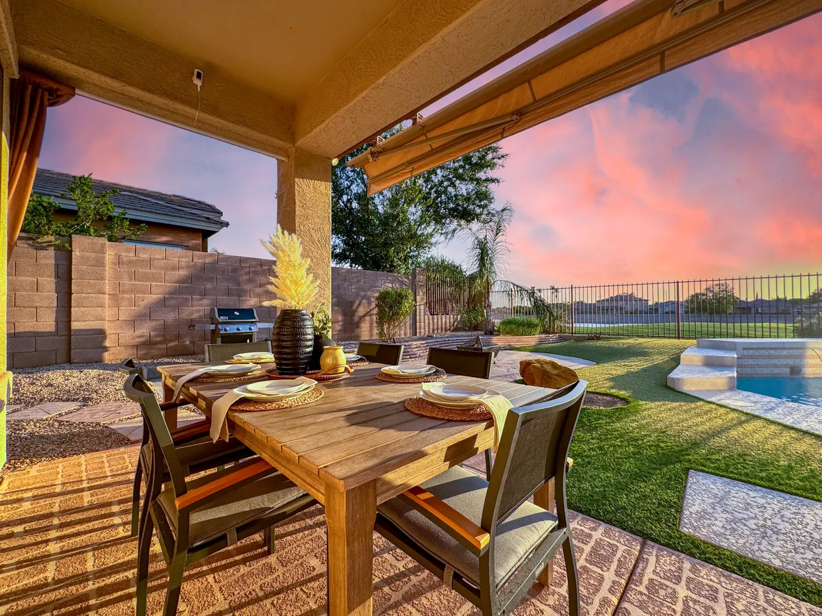 An outdoor living area featuring a large dining table, grill, shimmering pool, lush landscaping, and a warm sunset sky. The scene suggests an inviting space for outdoor meals, barbecues, and poolside relaxation, with a welcoming ambiance for gatherings.