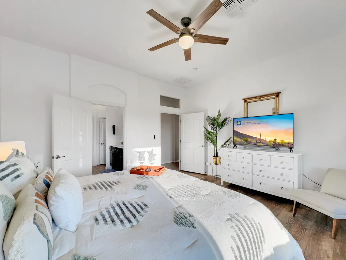 A serene bedroom featuring a bed with decorative pillows, a television set above a dresser, and a lush green plant. Sunlight streams into the room, creating a warm and welcoming atmosphere ideal for relaxation and movie nights.